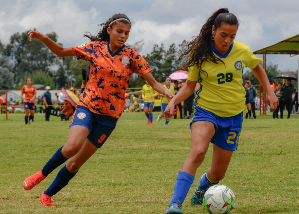 Two teenage girls actively competing in a soccer match on a grassy field, wearing sports uniforms.