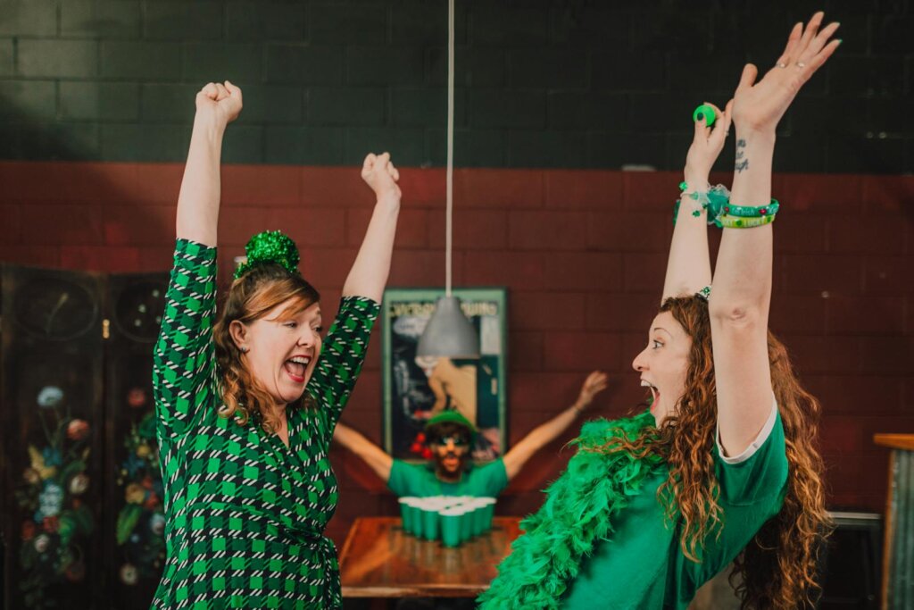 Two women celebrate St. Patrick's Day enthusiastically indoors with vibrant green attire and accessories.