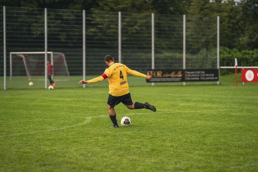 An amateur soccer player in yellow jersey kicks a ball on a grassy field during the day.